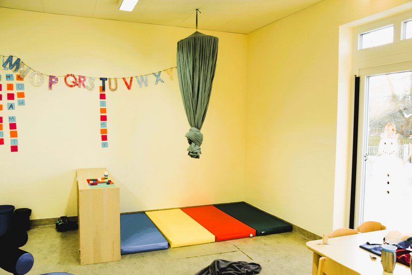 Bunte Matte auf dem Boden, darüber eine Stoffschaukel, an der Wand bunte Bilder