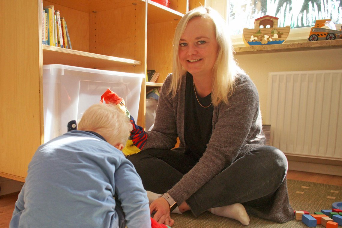Eine blonde Frau lächelt in die Kamera, von hinten von der Sonne beschienen, im Vordergrund ein Kind von hinten