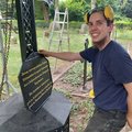 Junger Mann in Arbeitskleidung lächelt in die Kamera, hält schwarze Platte mit goldener Schrift