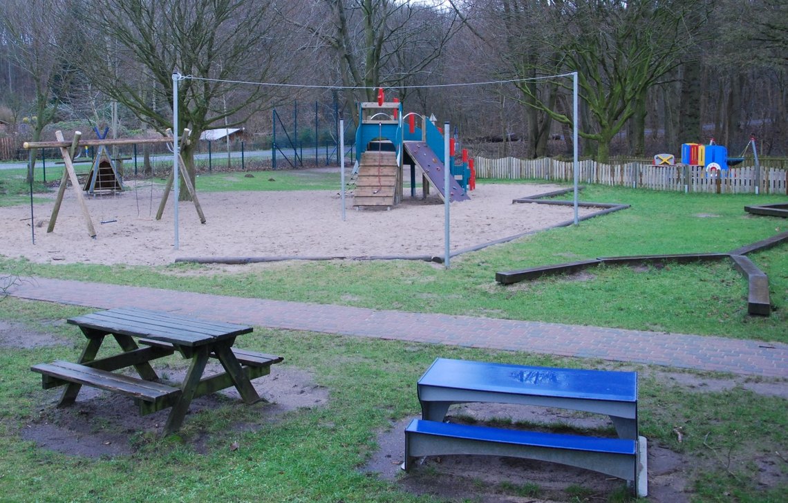 Vorne Sitzmöglichkeiten aus Holz und in blau, dahinter Schaukel, Balancierbalken und Klettermöglichkeiten